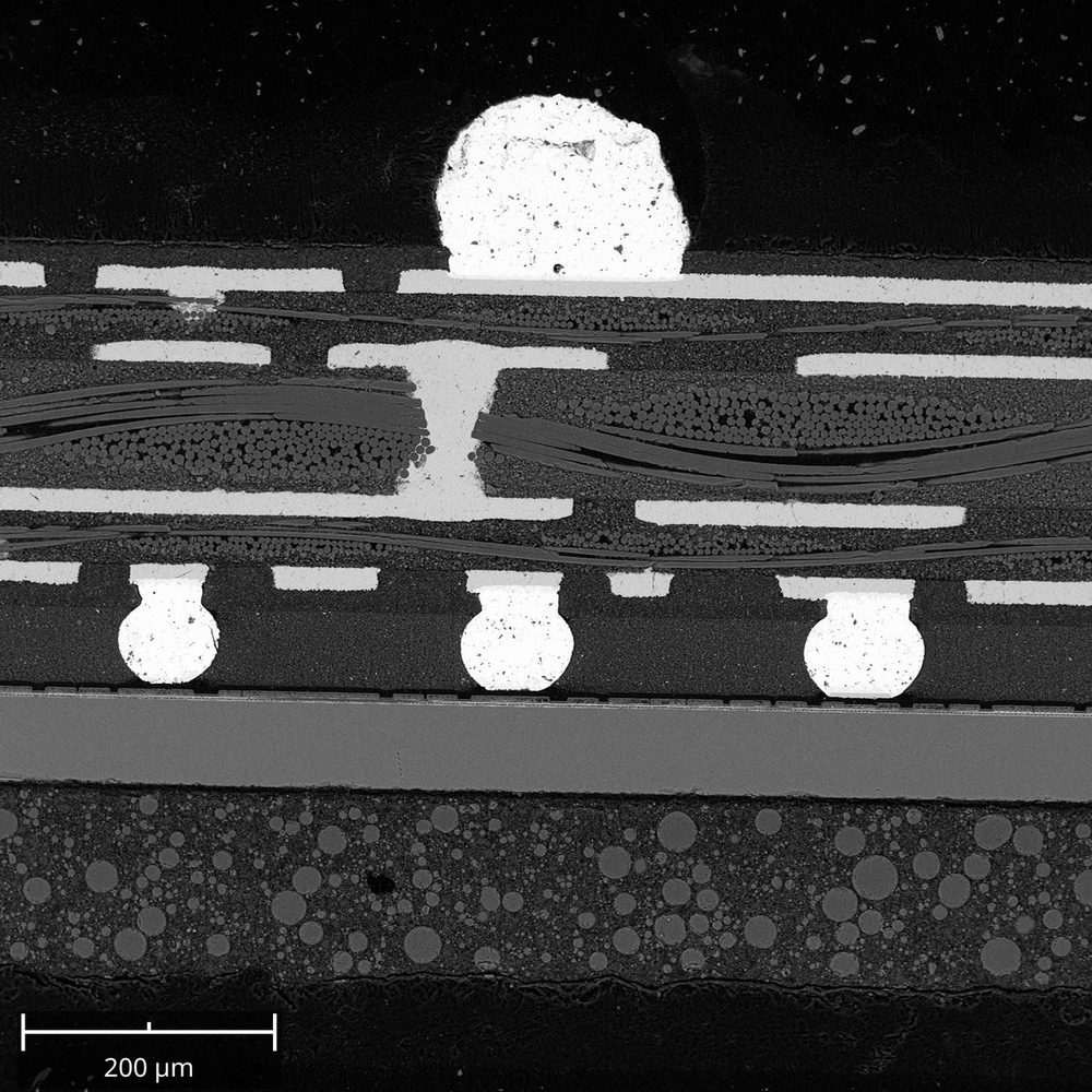 SEM image of a cross-section through an integrated circuit package: solder bumps, copper traces and a via, glass-fiber laminate, and particle-filled mold compound.